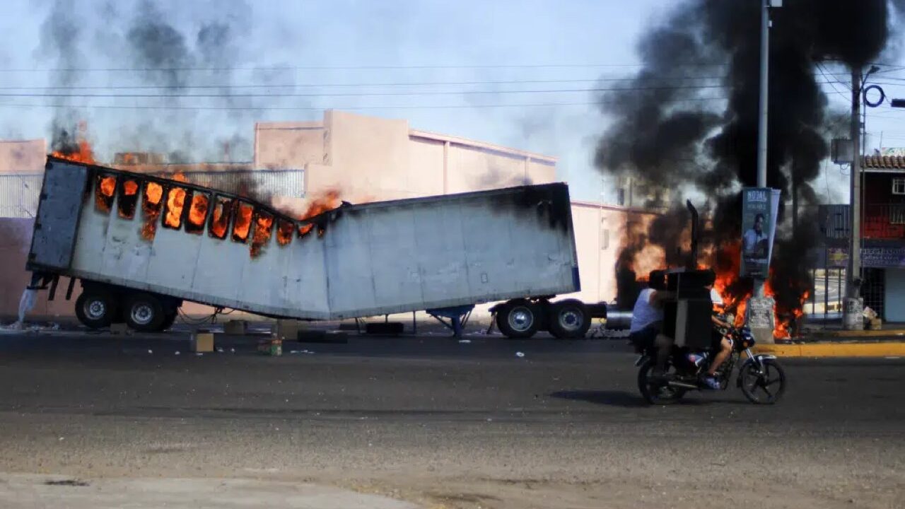 Dos hombres a bordo de una motocicleta pasan junto a un camión en llamas en las calles de Culiacán, en el estado de Sinaloa, el 5 de enero de 2023. (AP Foto/Martín Urista)